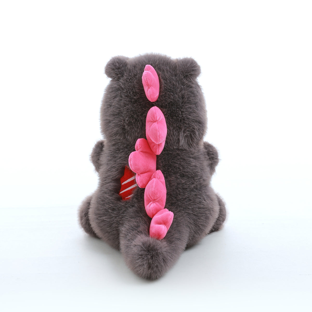 Grey kaiju baby plush toy viewed from behind, showing pink spikes along its back and tail on a white background.