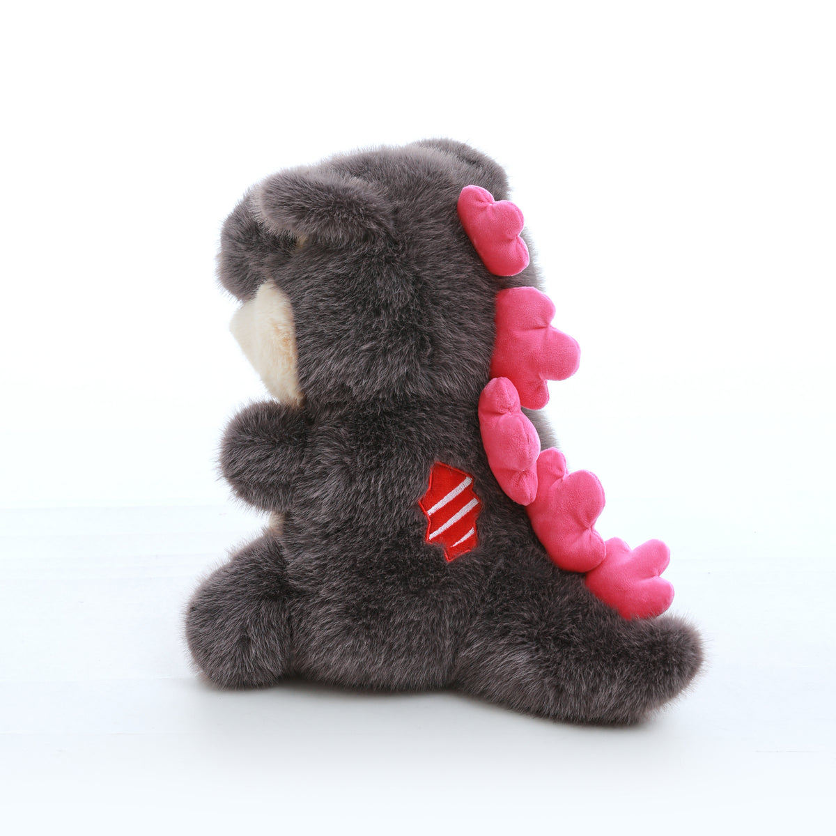 Grey kaiju baby plush toy with pink spikes along its back and tail, shown in a rear three-quarter view on a white background.